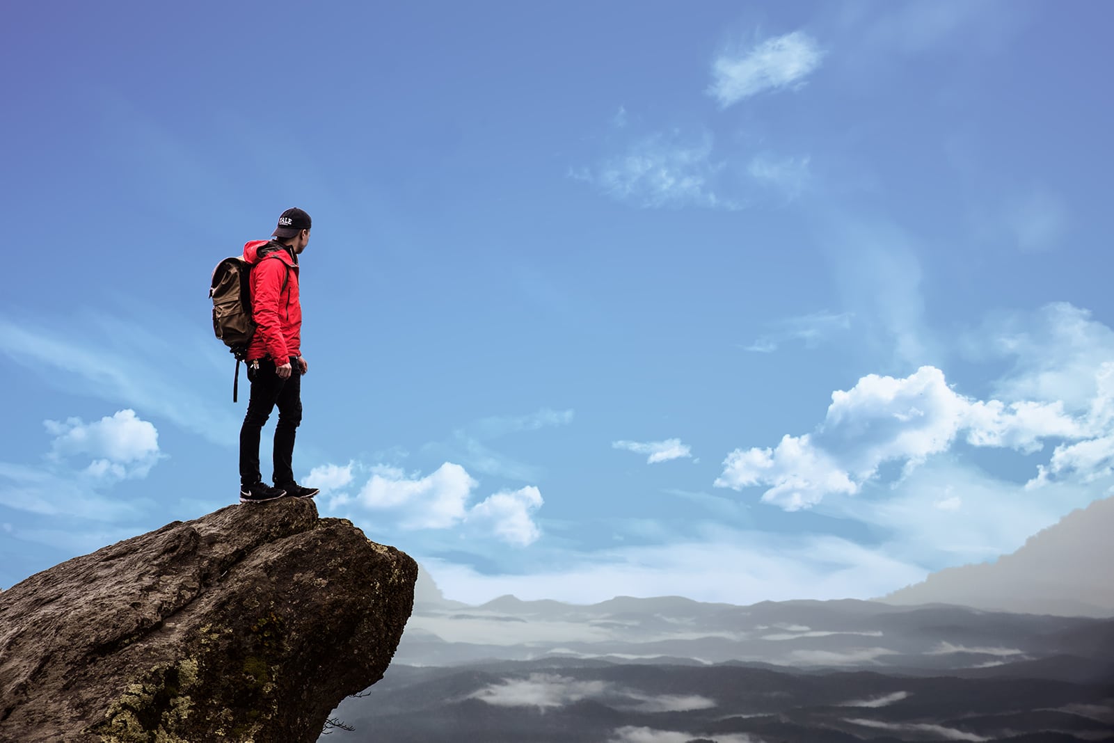 A hiker in a red jacket stands on a cliff with a backpack